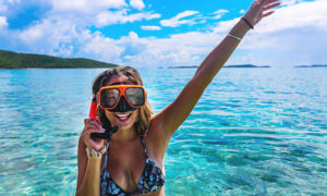 A woman with snorkeling gear smiling and waving as she emerges from the water.