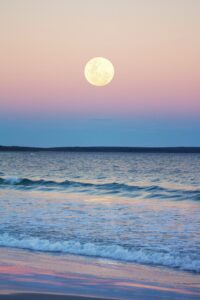 A full moon hanging over the ocean at sunset.